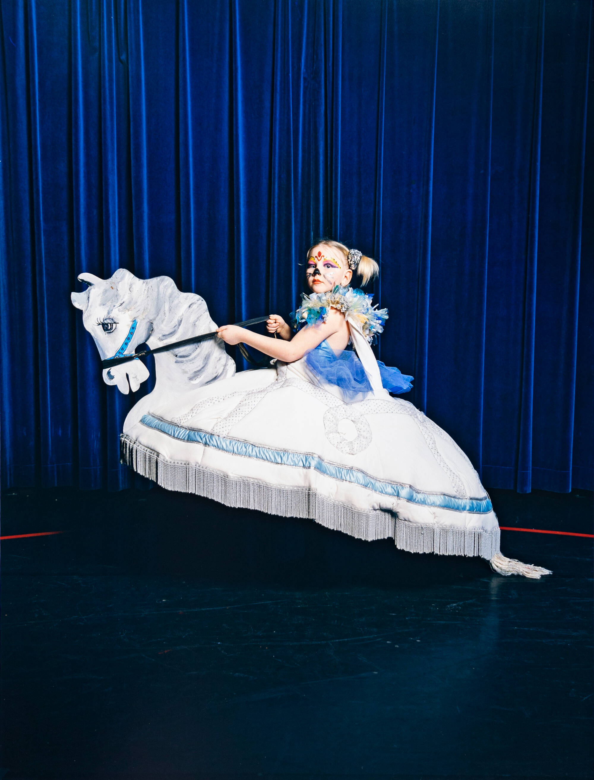 Girl in white dress with face paint, sitting on a pretend horse. In front of blue theater curtain.
